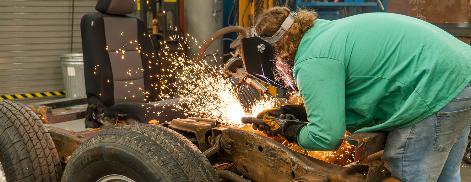 An Alamance Community College student uses a welding torch to cut or repair a vehicle frame, with bright sparks flying. The student is wearing protective gear, including gloves and a welding helmet, while working in an automotive lab setting.