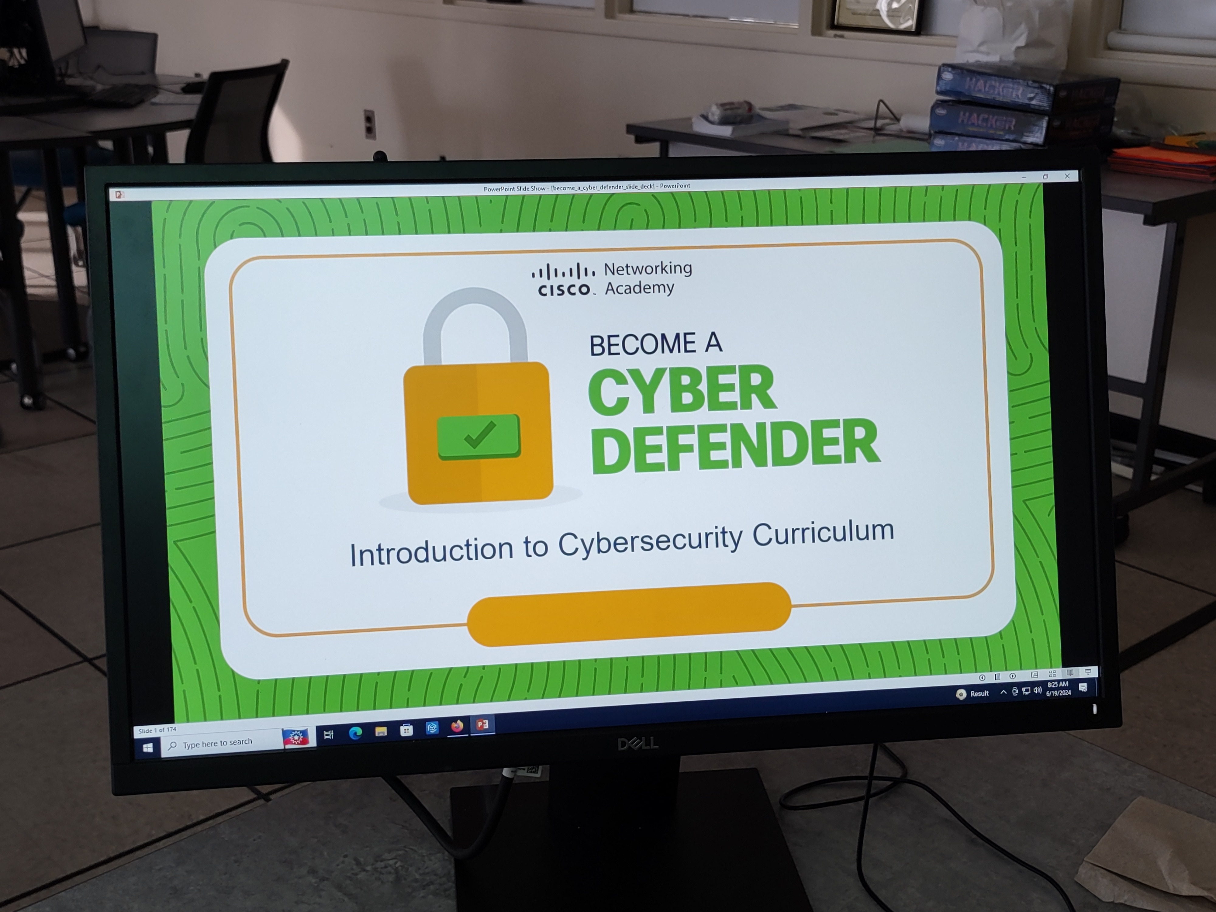 A computer monitor displaying a Cisco Networking Academy presentation slide titled 'Become a Cyber Defender: Introduction to Cybersecurity Curriculum.' The slide features a green and white design with a padlock graphic. The background shows a classroom setting with tables, chairs, and cybersecurity-related materials