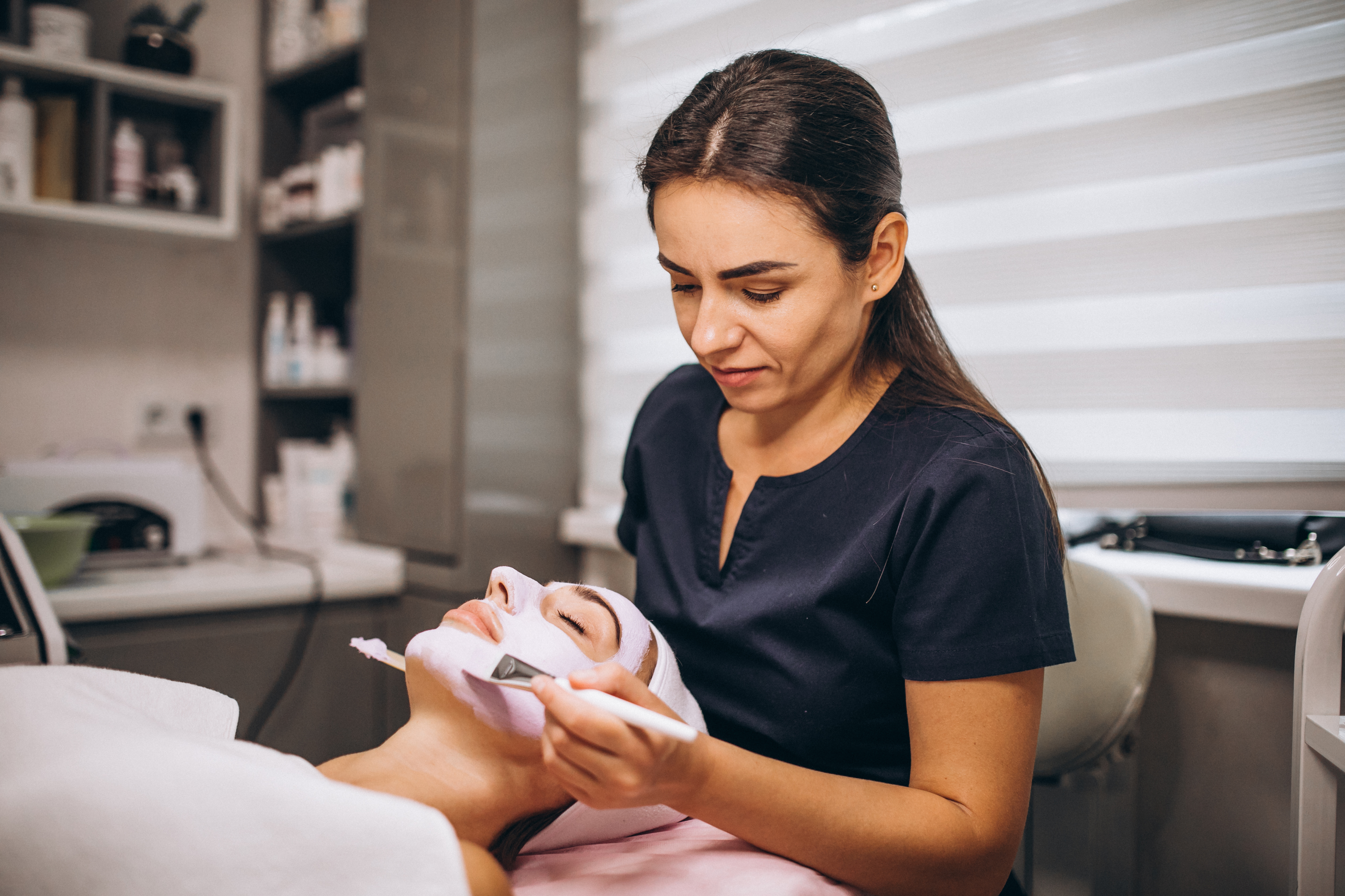 Cosmetology student applying a facial mask to a client at a beauty salon, demonstrating skincare techniques in a hands-on learning environment.
