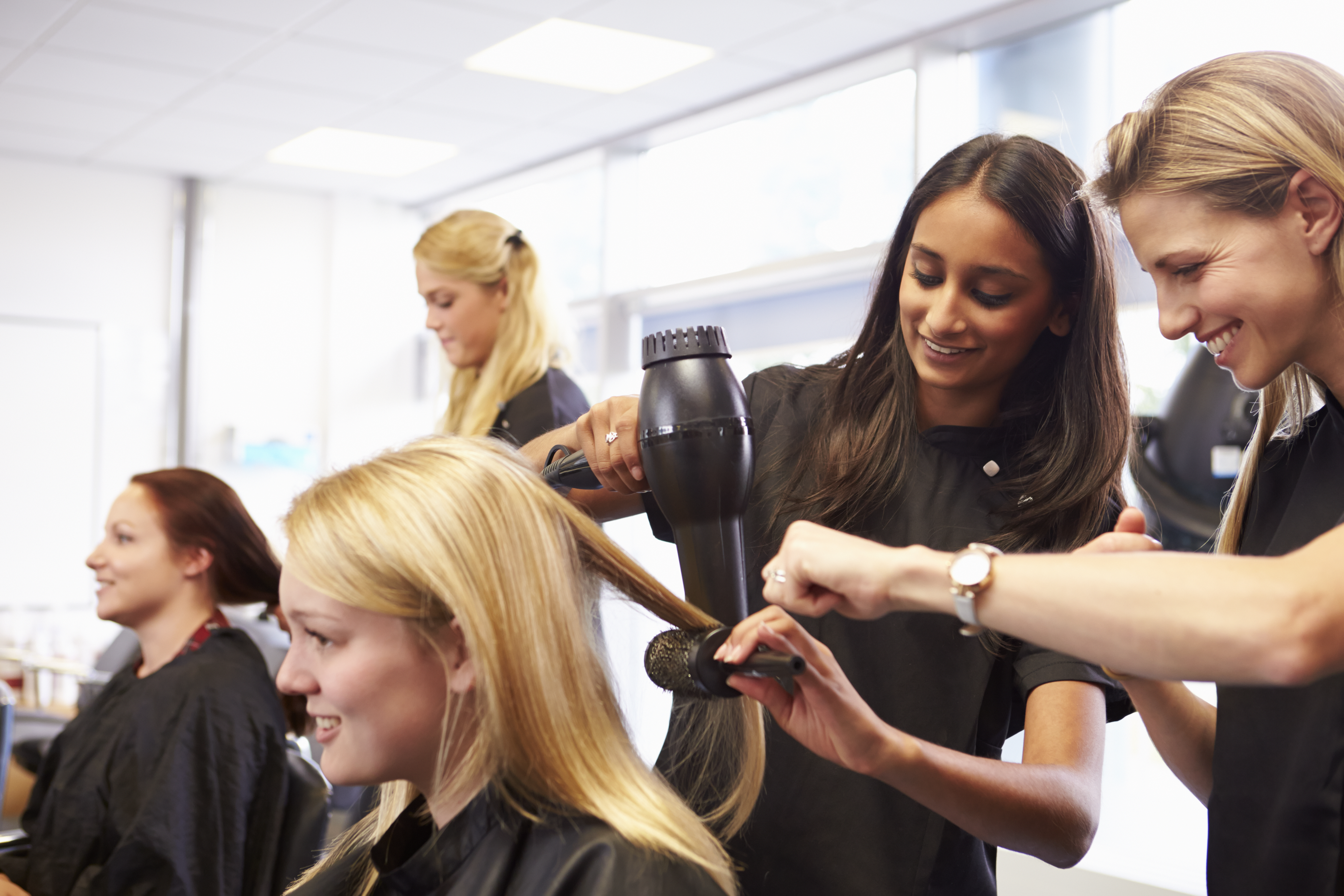 Cosmetology instructor guiding a student as she blow-dries a client's hair, with other students practicing hair styling techniques in the background.