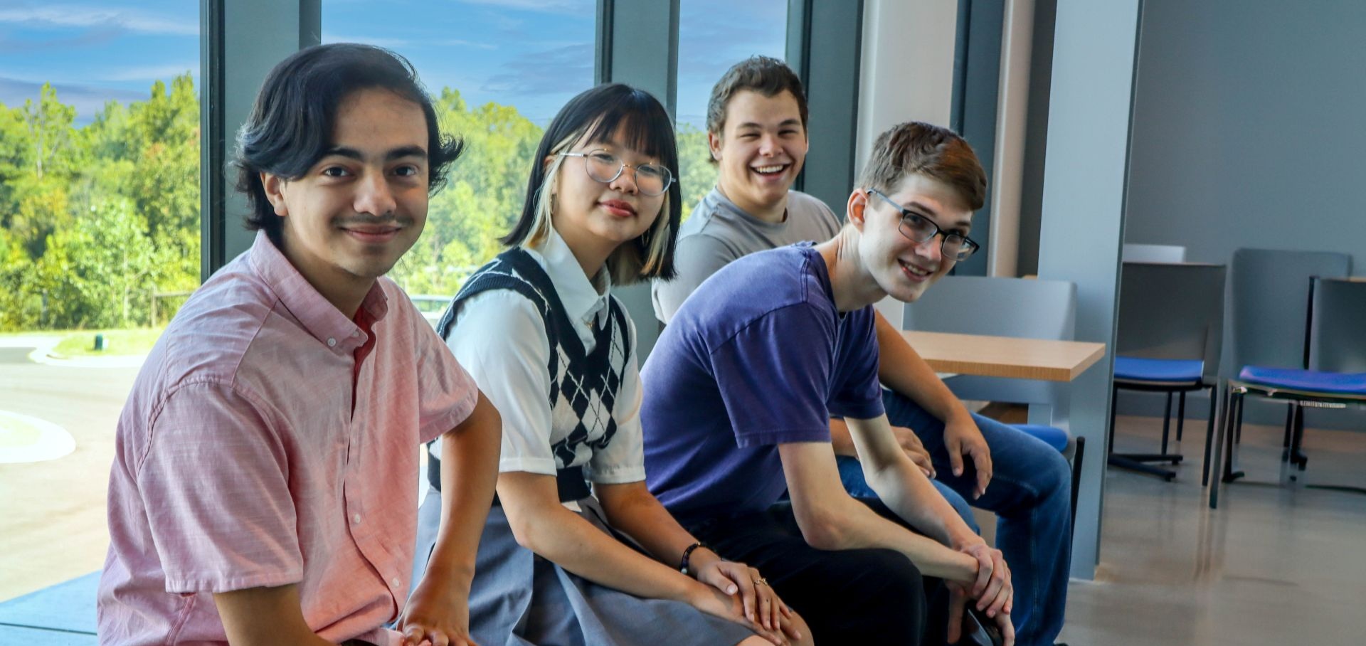 Four Alamance Community College students sit together by large windows in a modern campus building. They are smiling and casually dressed, with a scenic view of trees and blue sky in the background, reflecting a welcoming and inclusive learning environment.