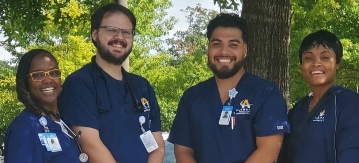Four Alamance Community College Associate in Nursing (ADN) students wearing navy blue scrubs with ID badges, standing outdoors in front of trees and smiling. The group includes two women and two men, showcasing a diverse and professional team of future nurses.