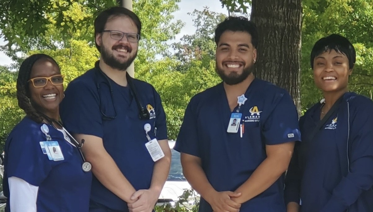 Four Alamance Community College Associate in Nursing (ADN) students wearing navy blue scrubs with ID badges, standing outdoors in front of trees and smiling. The group includes two women and two men, showcasing a diverse and professional team of future nurses.