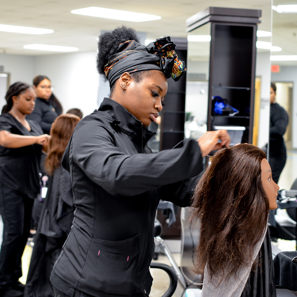 An Alamance Community College cosmetology student styles a mannequin head in a salon classroom. Other students practice hair techniques in the background, creating a hands-on learning environment for hair design and cosmetology skills.