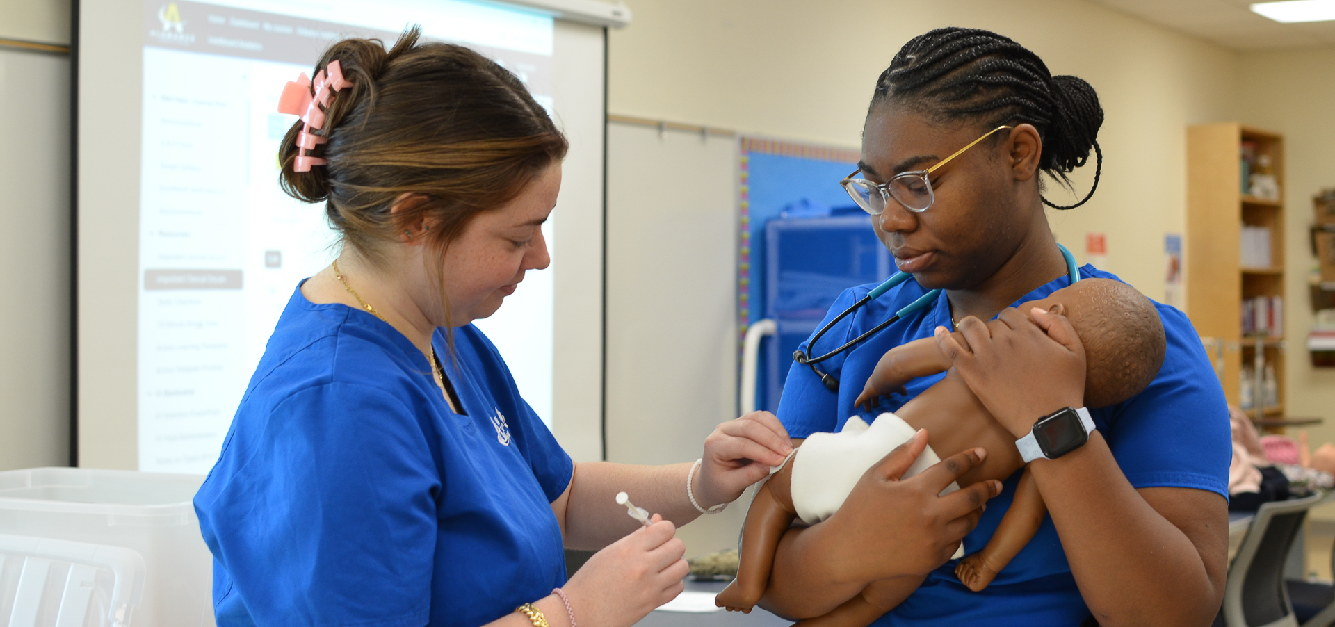 Two Alamance Community College nursing students practice pediatric care using an infant mannequin. One student gently holds the mannequin while the other prepares to administer care with a syringe and cotton swab in a clinical lab setting.