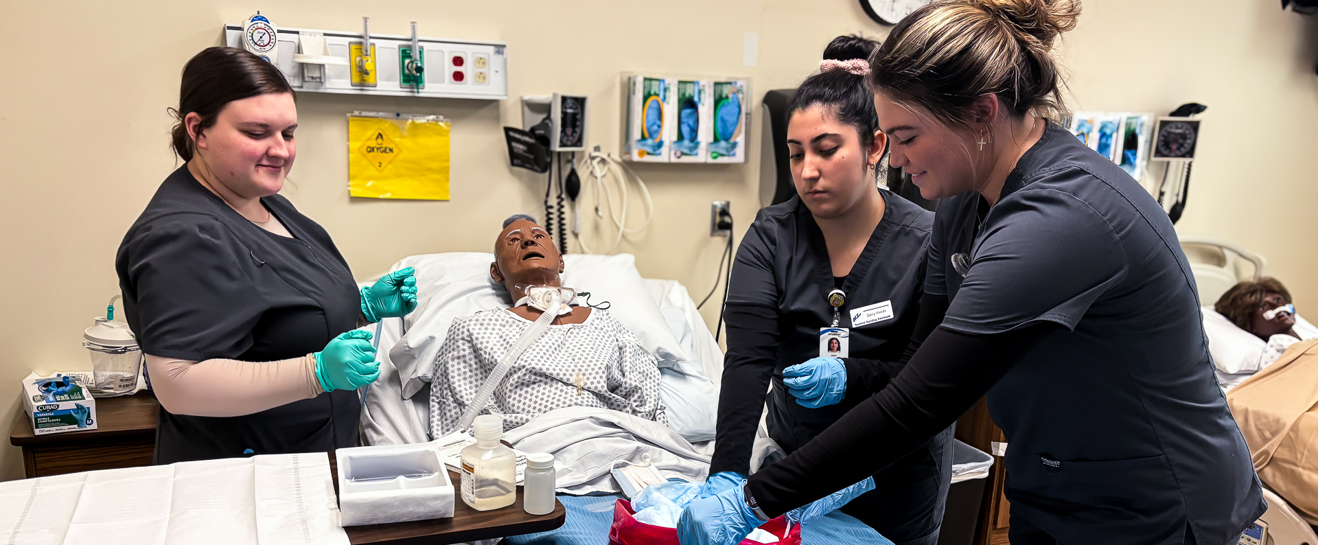 Alamance Community College nursing students practice clinical skills on a medical simulation mannequin in a lab setting. The students are wearing gloves and working collaboratively around a hospital bed equipped with realistic medical tools and monitors.