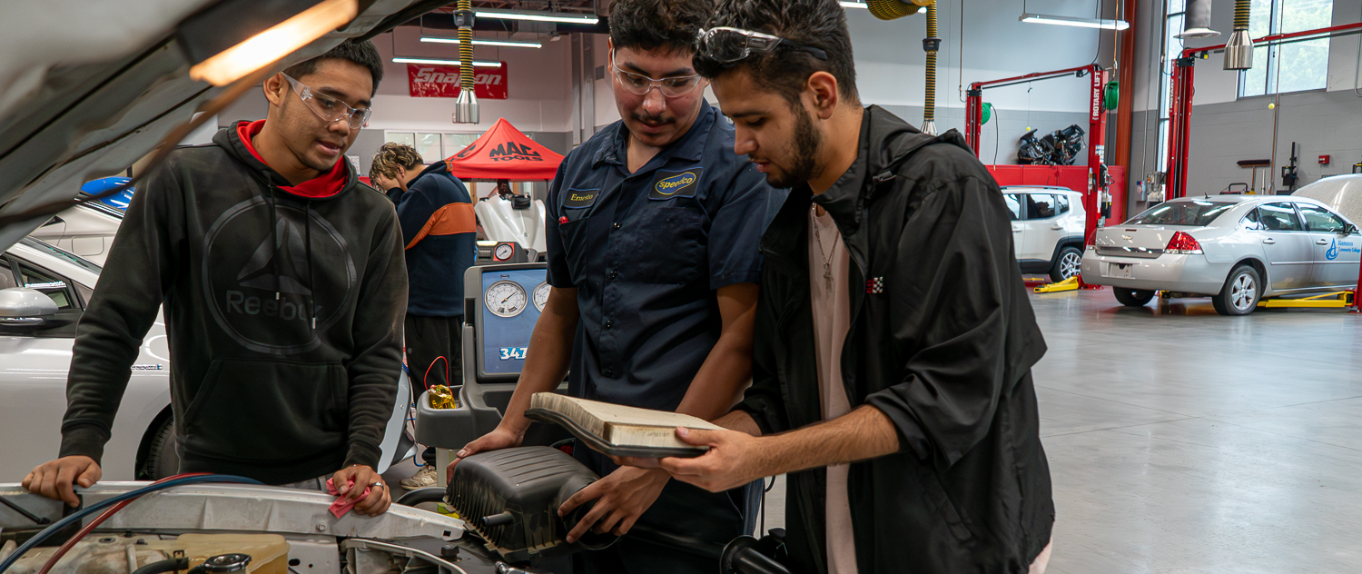 Three Alamance Community College automotive students work together under the hood of a vehicle in a campus garage. They are examining an air filter while wearing safety glasses, with various diagnostic equipment and vehicles visible in the background.