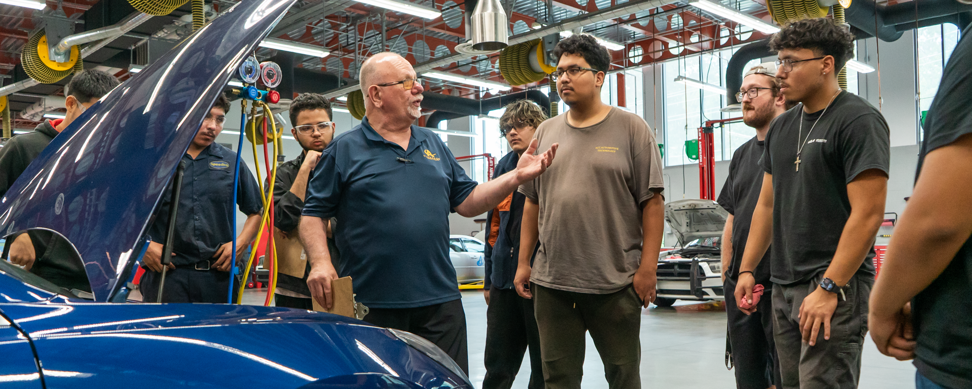ACC automotive instructor teaching a group of students gathered around a car with the hood open in a college auto technology lab.