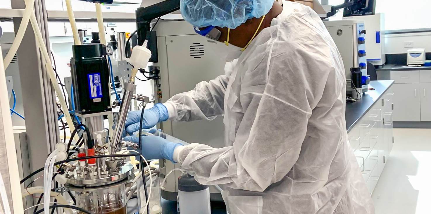 Laboratory setting with an Alamance Community College student wearing white lab coat, hair net, and mask working at biosafety cabinets. They are handling equipment and materials in a clean, well-lit environment. The lab features advanced technology and equipment, highlighting a professional biotechnology facility