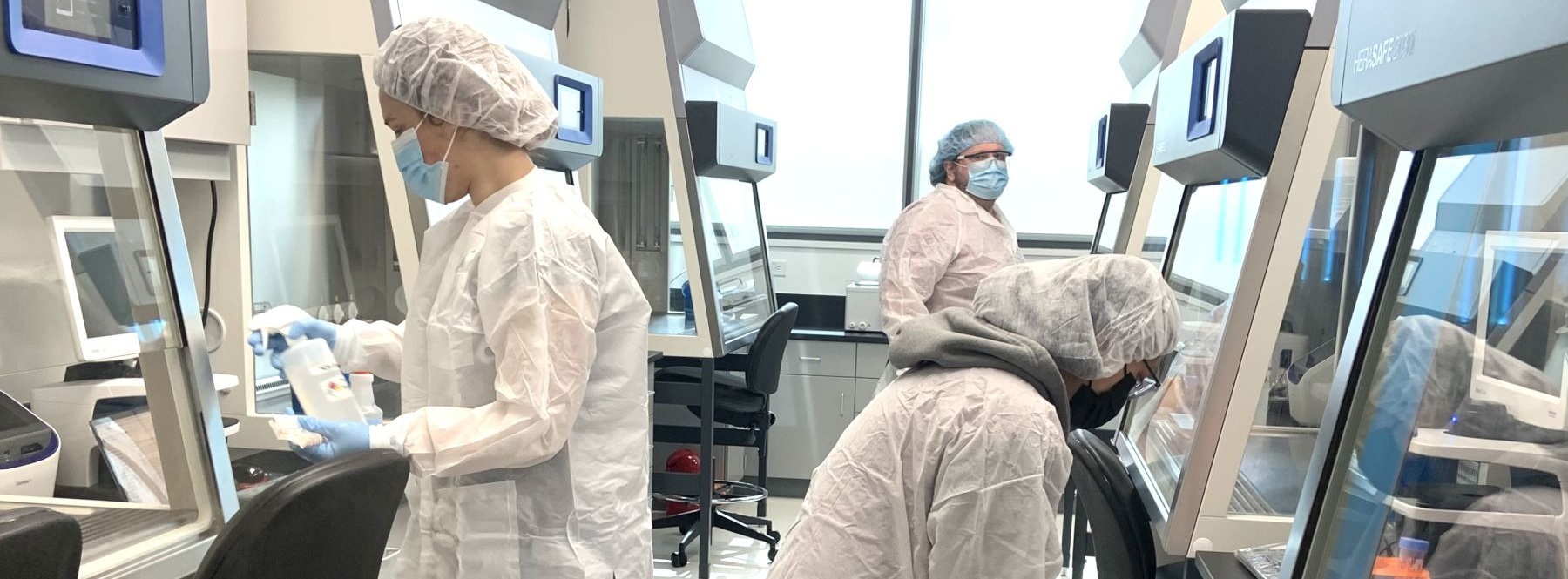 Laboratory setting with three Alamance Community College students wearing white lab coats, hair nets, and masks working at biosafety cabinets. They are handling equipment and materials in a clean, well-lit environment. The lab features advanced technology and equipment, highlighting a professional biotechnology facility