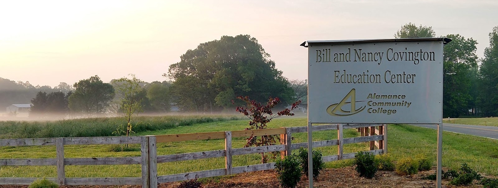 The Bill and Nancy Covington Education Center at Alamance Community College. The image shows a scenic view of a wooden fence enclosing a green field with trees and light morning mist in the background. A sign prominently displays the center's name along with the Alamance Community College logo, emphasizing the rural and serene setting of the Covington farm.