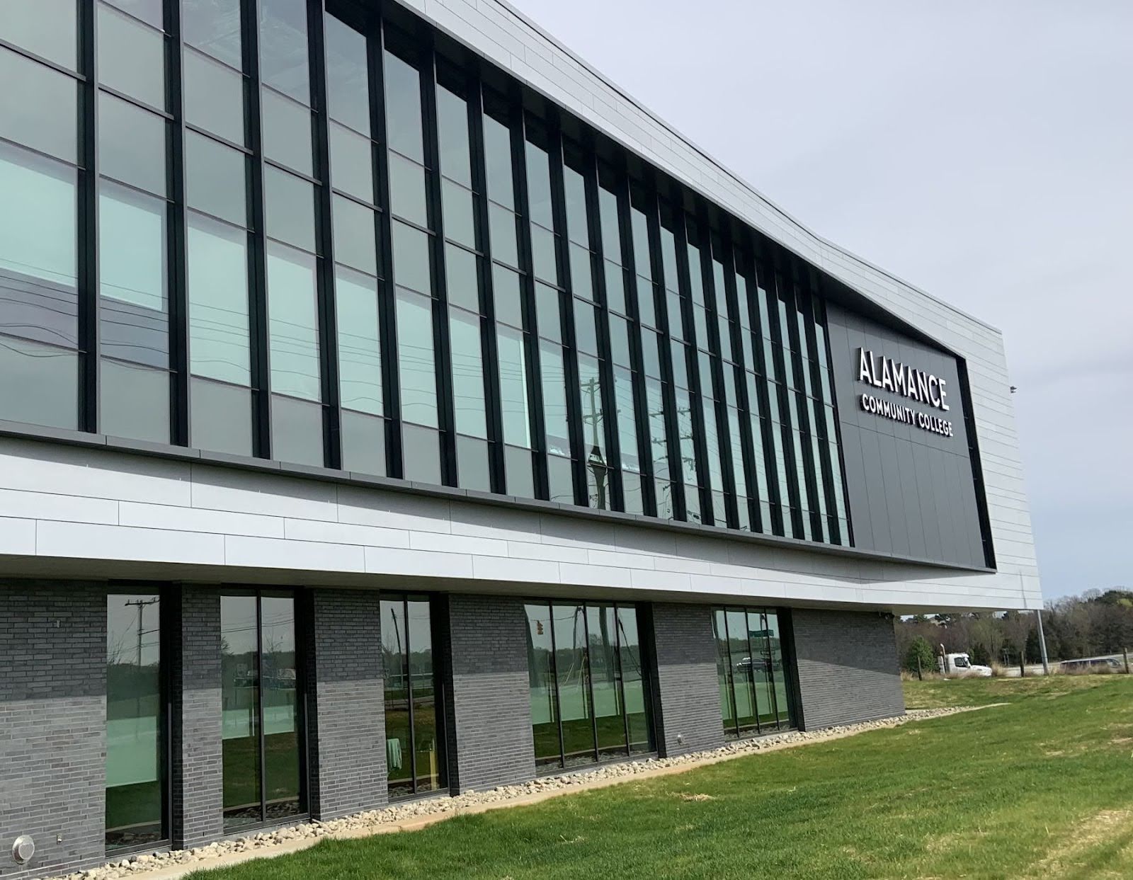 The Biotechnology Center of Excellence building at Alamance Community College. The modern structure features large glass windows and sleek architectural design, set against a backdrop of green grass and an overcast sky. The Alamance Community College logo is prominently displayed on the building's facade.