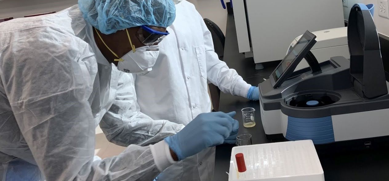 Two Alamance Community College Biopharmaceutical Technology students working in a laboratory. Both are wearing protective gear, including lab coats, gloves, masks, and hair covers, while conducting experiments. One student is handling a beaker near advanced lab equipment, emphasizing hands-on training in biopharmaceutical processes