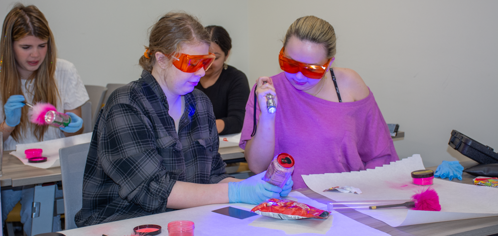 ACC students wearing safety goggles examining fingerprint evidence with UV flashlights during a forensic science class