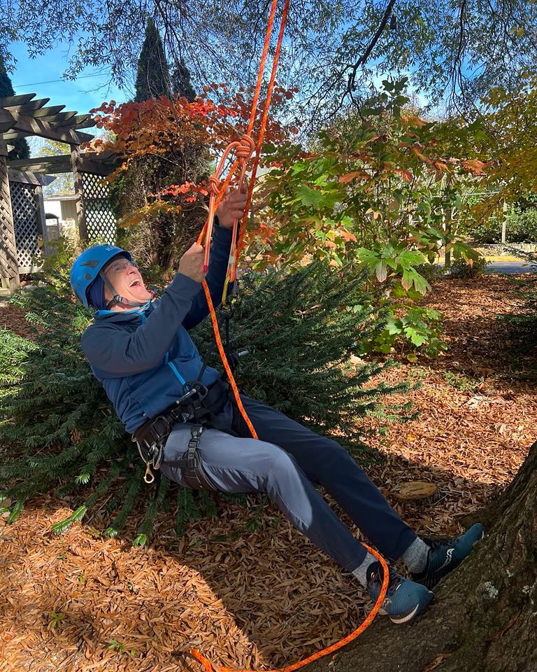 An acc student wearing a blue helmet and harness practicing tree climbing with bright orange ropes outdoors. The individual is smiling while holding and adjusting the rope system, surrounded by trees and autumn foliage in a natural setting.