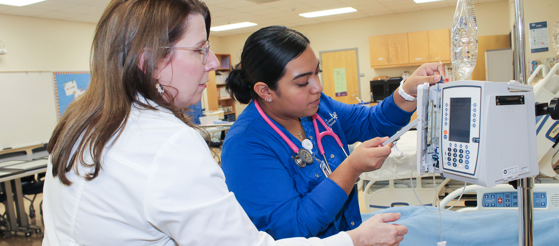 An Alamance Community College nursing student practices using an IV infusion pump under the guidance of an instructor. The student is wearing blue scrubs and a pink stethoscope in a simulated healthcare lab environment.