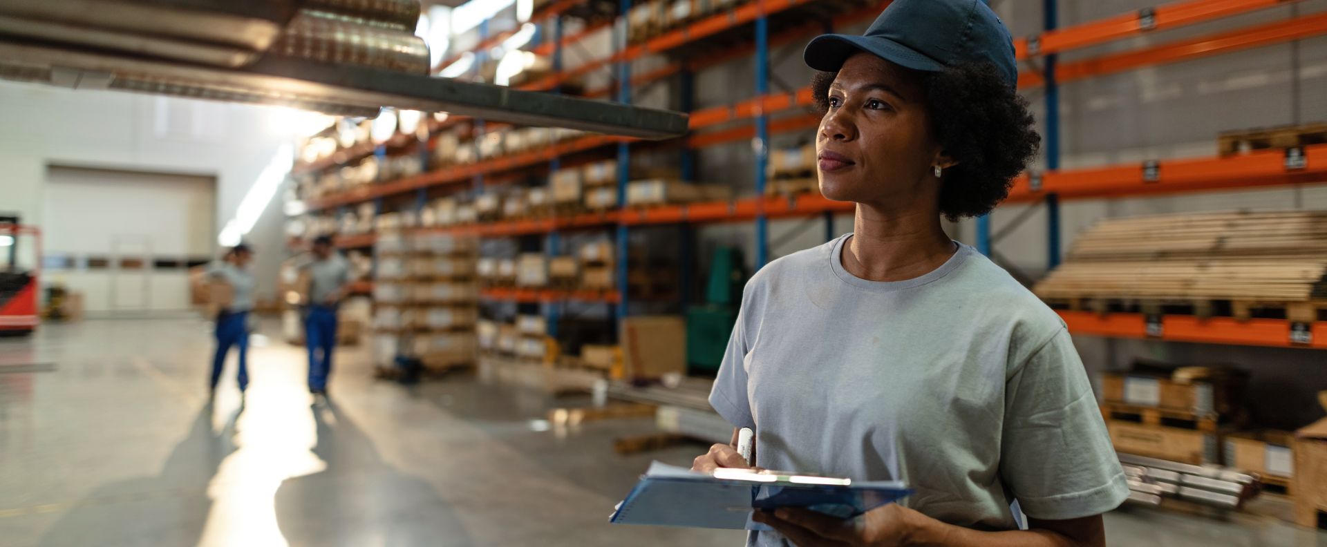 Warehouse worker holding a clipboard and looking ahead in a large storage facility with shelves of inventory in the background.