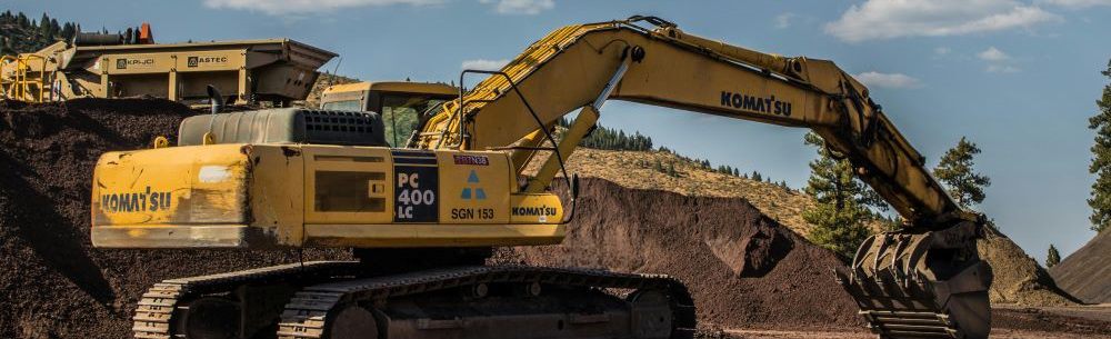 A large yellow excavator at a construction site, positioned on dirt with a mound of soil behind it. The excavator's arm is extended with a bucket attachment, and industrial equipment and machinery are visible in the background under a partly cloudy sky.