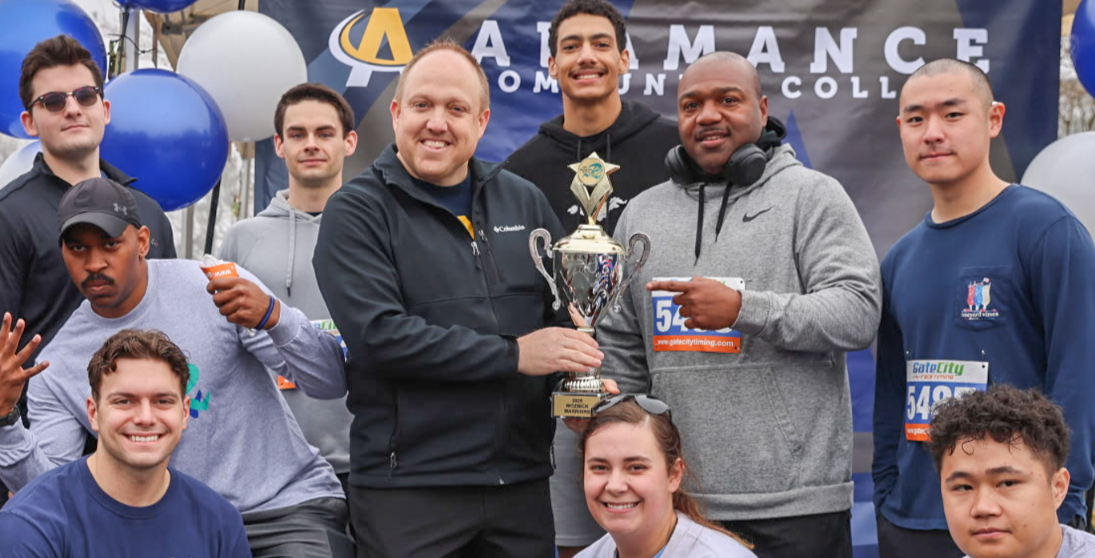 Group of Alamance Community College students and staff celebrating with a trophy at an outdoor race event, surrounded by balloons and a branded backdrop.