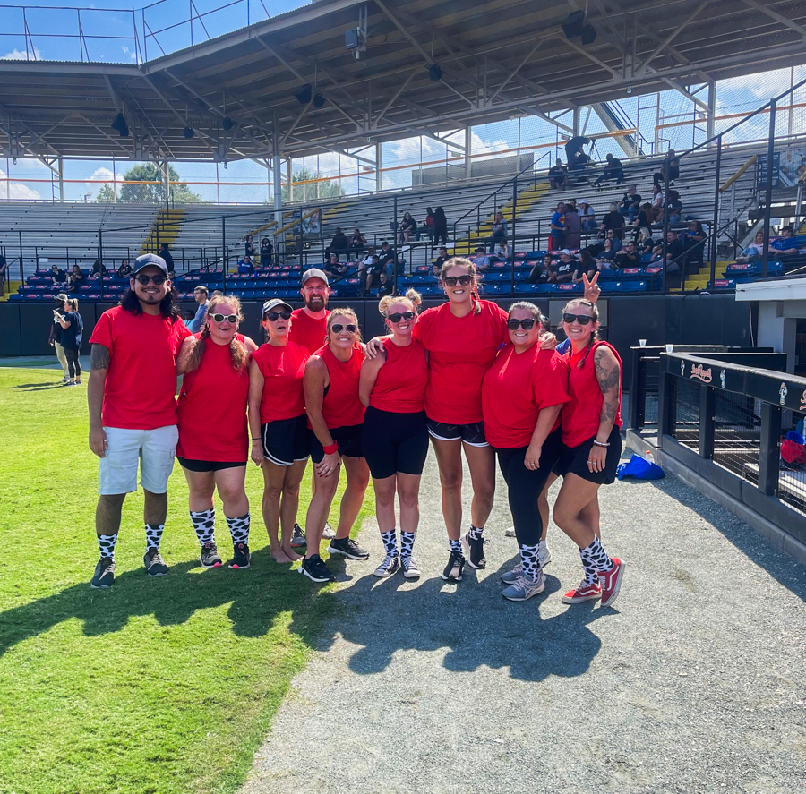 Group of Alamance Community College staff or students in matching red shirts posing together on a baseball field during a sunny outdoor event, with stadium seating and spectators in the background.