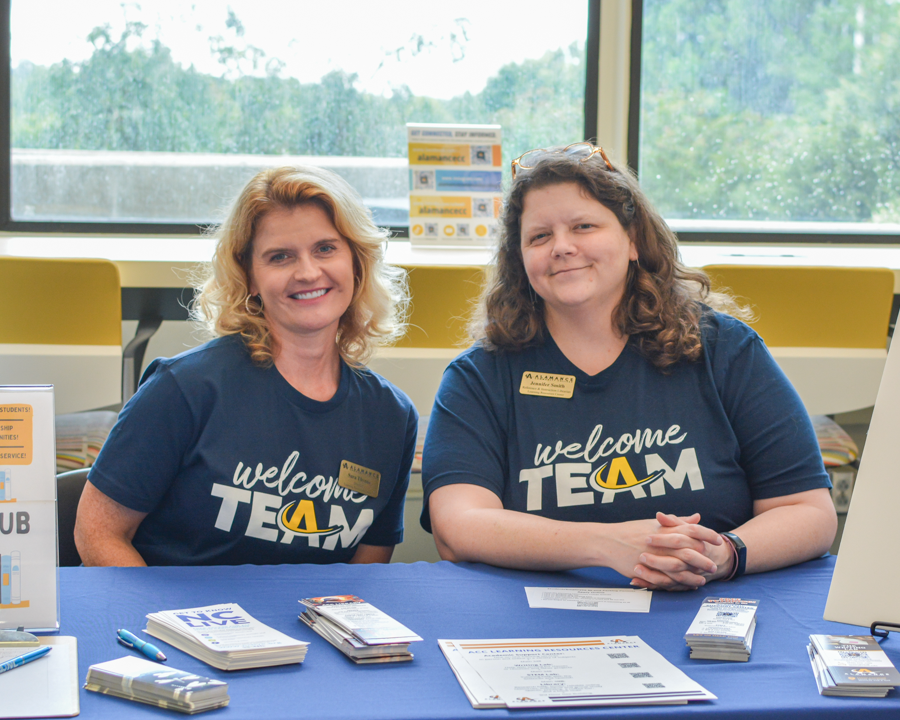 Two ACC employees smiling and collaborating at a table with a laptop and notepad in a bright, window-lit meeting space.