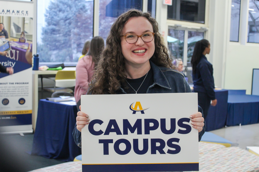Student recruiter holding a 'Campus Tours' sign inside Alamance Community College during a recruitment event, with booths and attendees visible in the background.