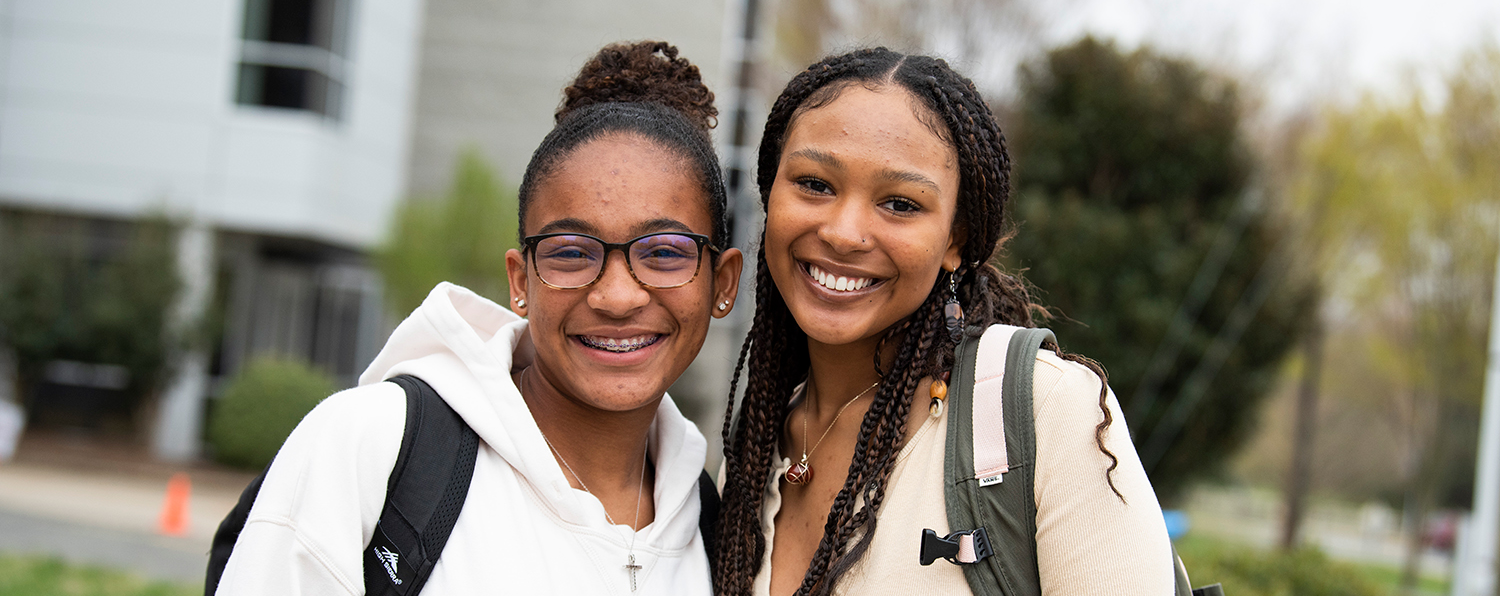 mentor program Two smiling students standing outdoors on campus with backpacks, posing together in front of a campus building.