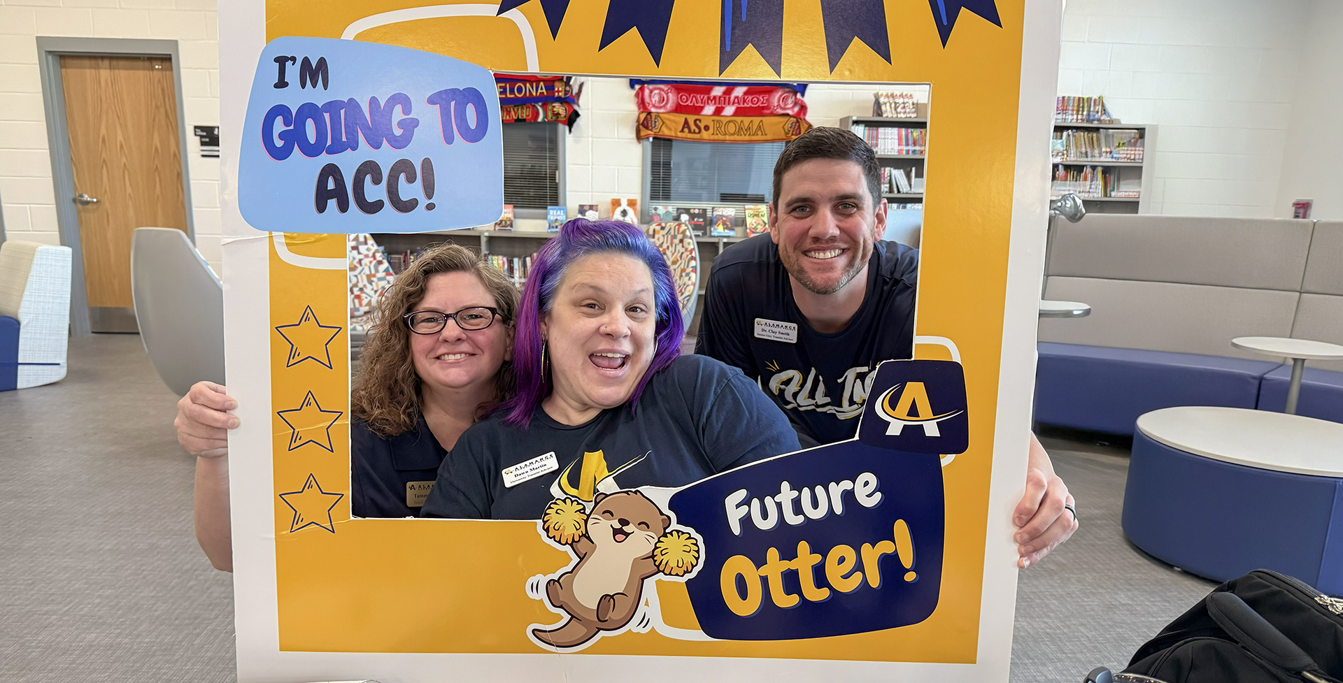 Three advising staff members smiling and posing behind a photo frame sign that reads 'I’m going to ACC! Future Otter!' inside a campus library space.