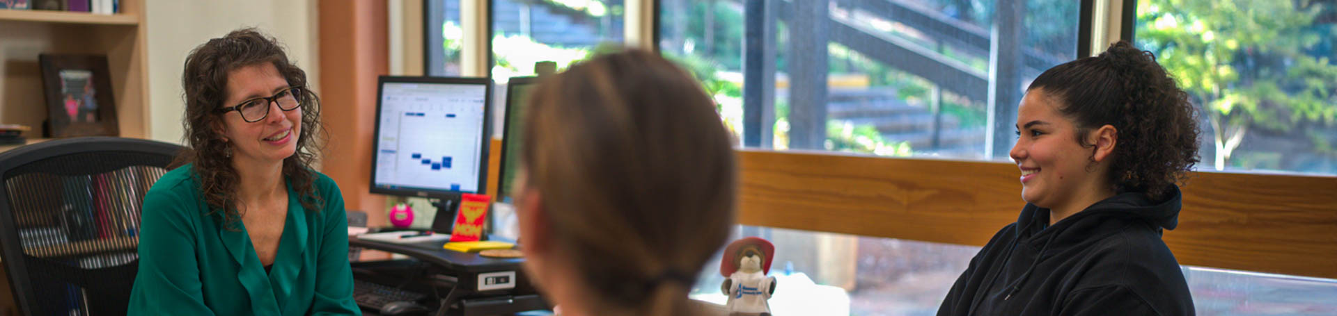 An Alamance Community College staff member meets with two students in her office, offering guidance and support. The space is bright and welcoming, with natural light coming through the windows and personalized decor on the shelves and desk.