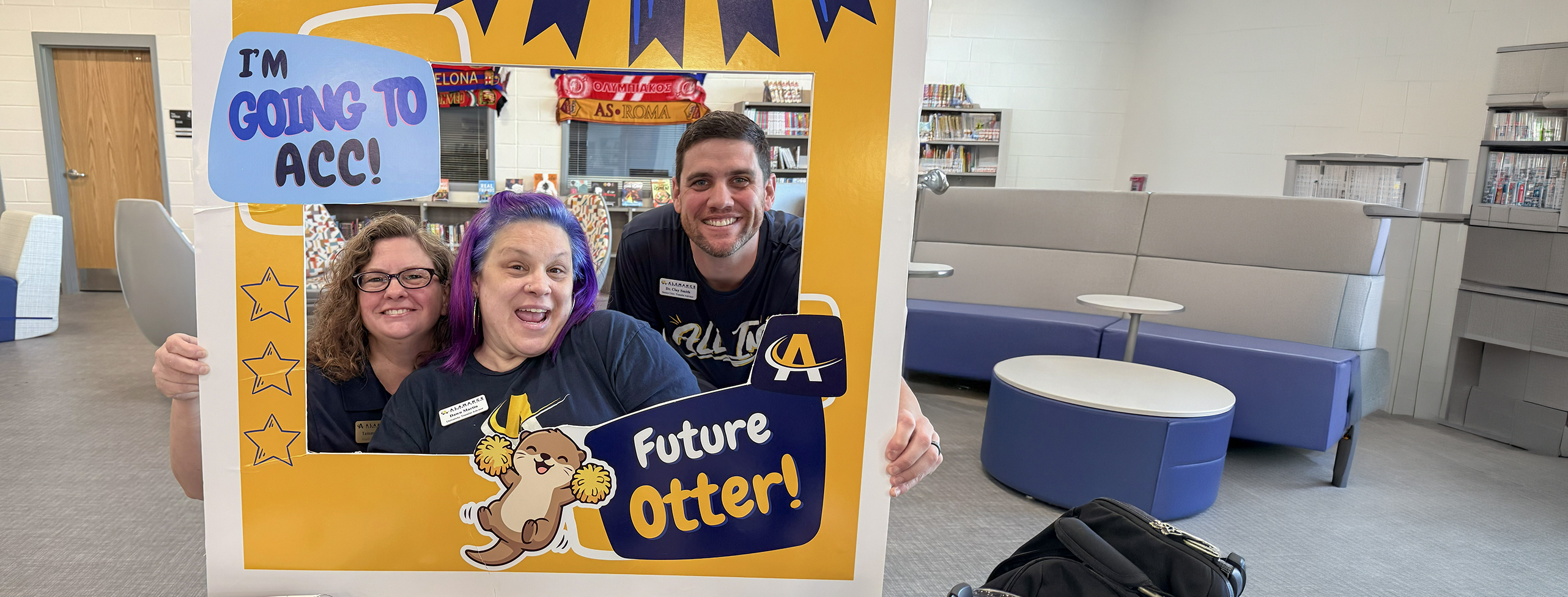 Three staff members smiling and posing behind a photo frame sign that reads 'I’m going to ACC! Future Otter!' inside a campus library space.