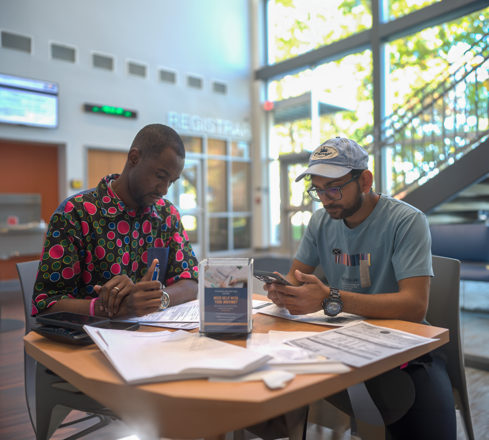 Two students seated at a table in a bright, modern campus building, working on paperwork and using a smartphone. One student wears a colorful polka-dot shirt and is focused on filling out forms, while the other, in a light blue shirt and cap, looks at his phone. The background includes large windows, a registration sign, and a staircase, creating an open, inviting atmosphere