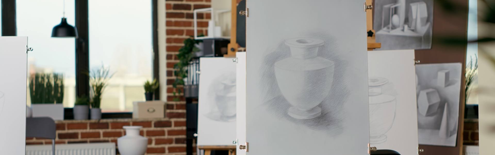Drawing Art classroom with easels displaying pencil drawings of still-life objects, including a shaded vase, in a well-lit studio with brick walls and windows.